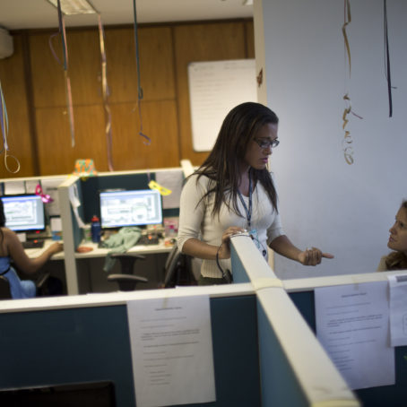 A woman in white and spectacles, wearing a name tag lanyard, talking to another woman who is sitting.