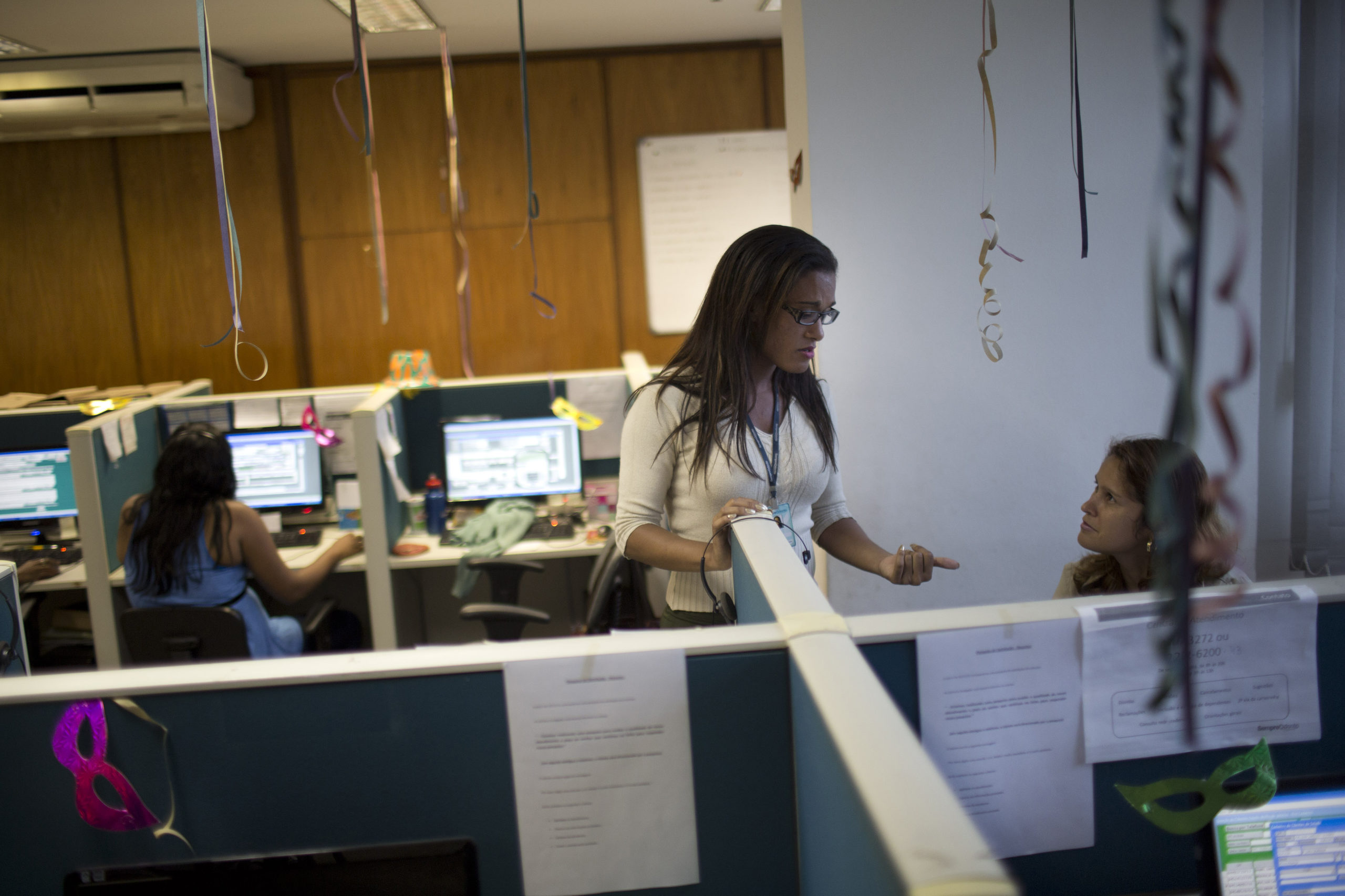 A woman in white and spectacles, wearing a name tag lanyard, talking to another woman who is sitting.