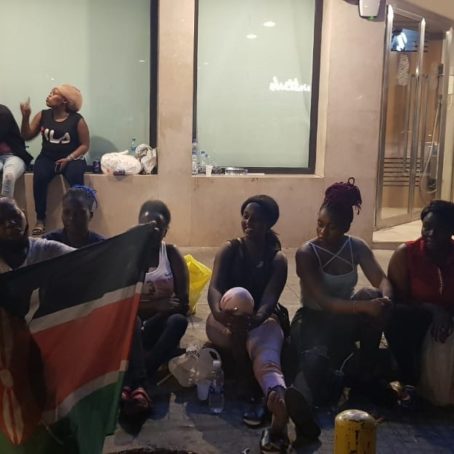 A large group of women sit on the pavement; a Kenyan flag held against the tree.