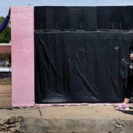 photo of a woman standing next to a building with another person walking past