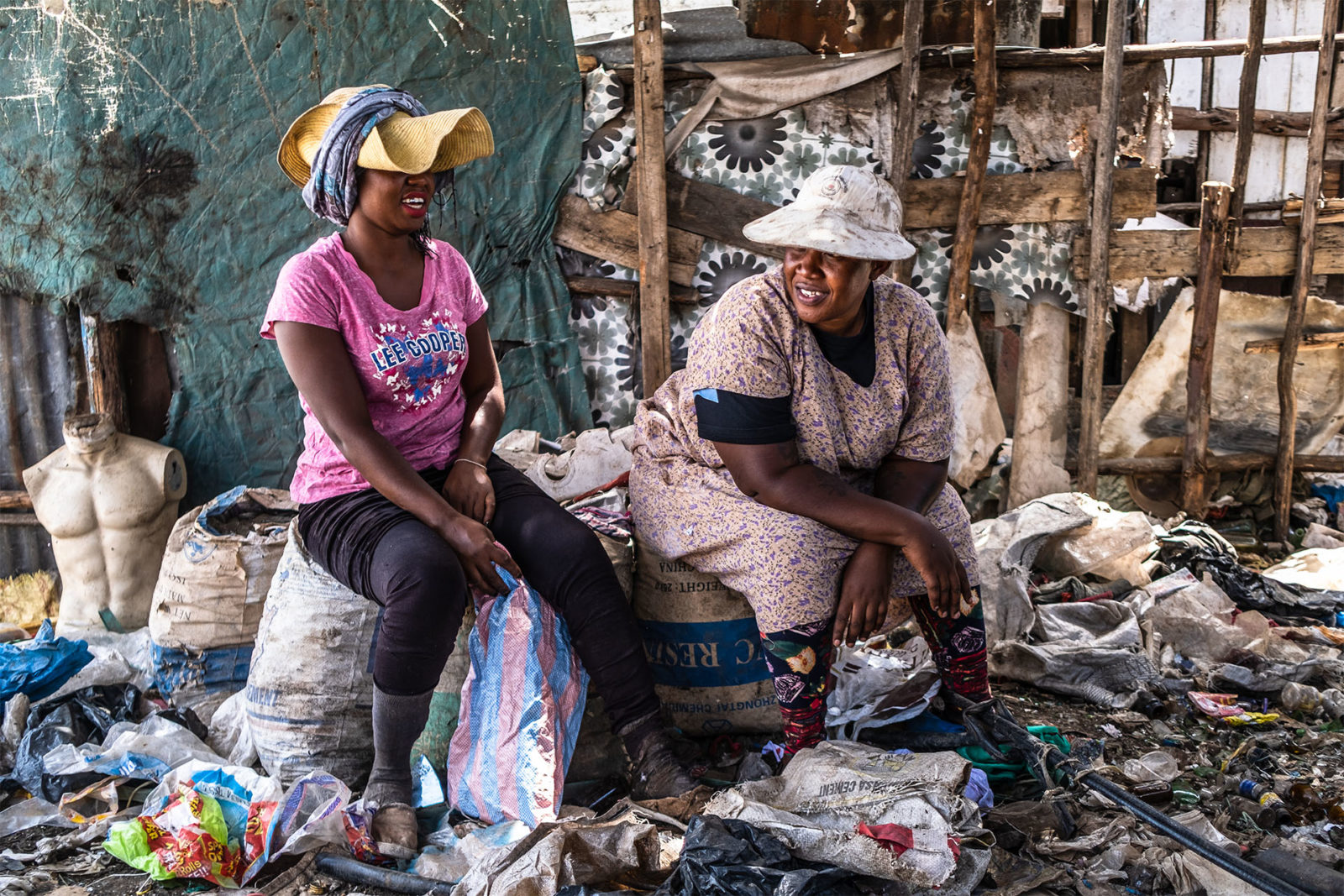 A photo of two women laughing, sitting on trash bags at a dumpsite