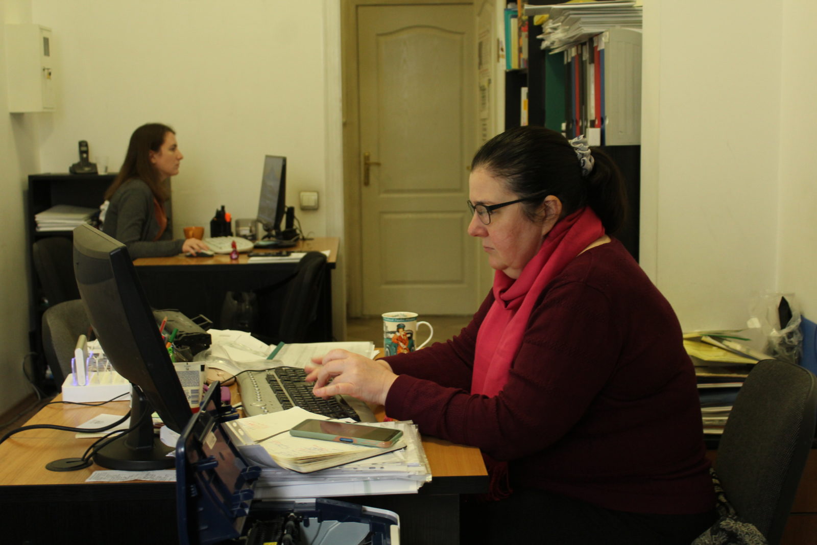 Two women sit across from each other at their desks typing on computers.