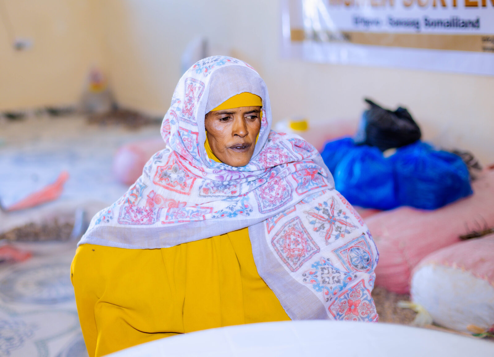 A woman wearing a head scarf and a yellow outfit sits at a white table.
