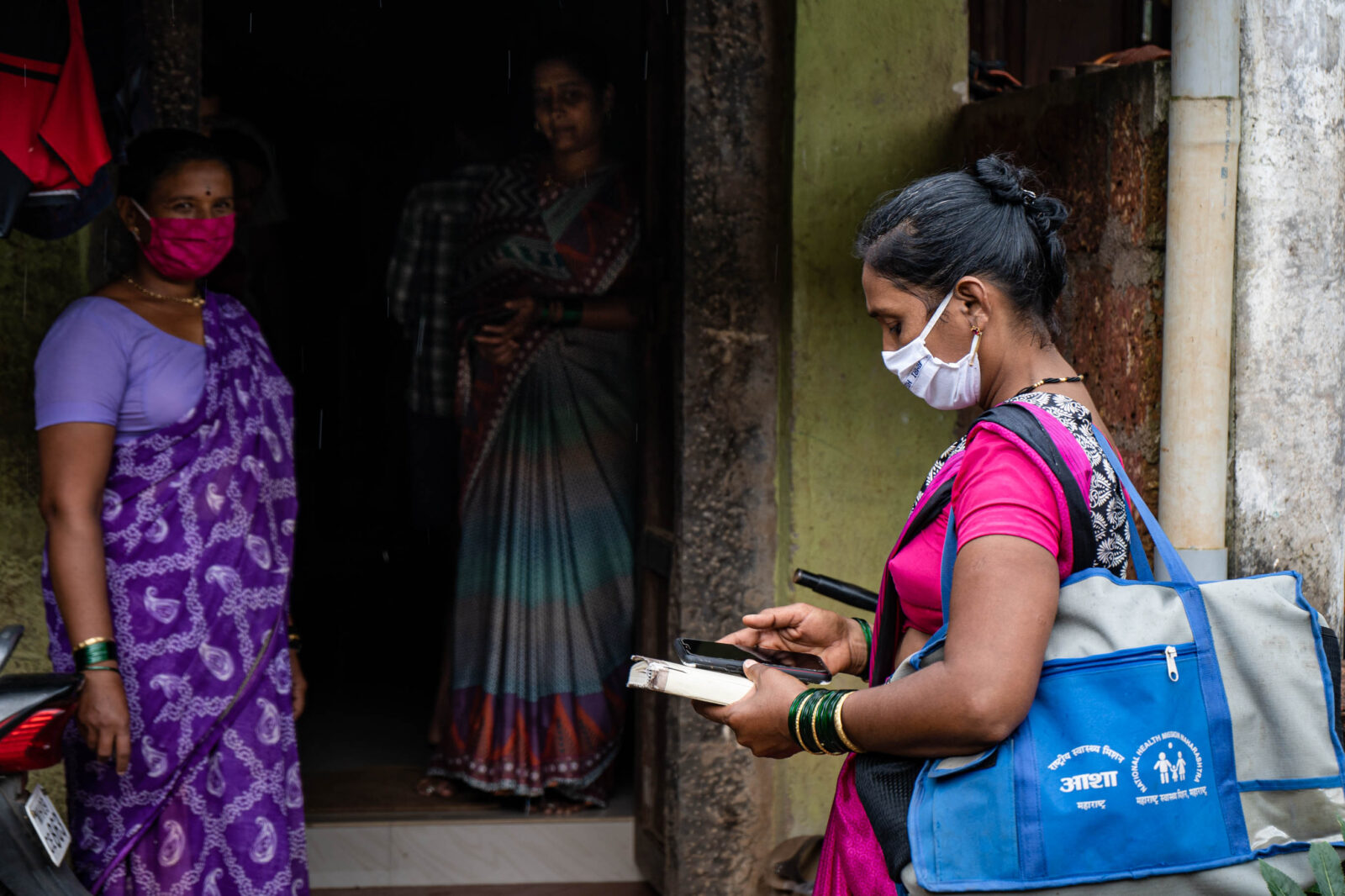 A woman stands outside a door carrying a blue tote bag and holding a sheet of paper in front of two women.