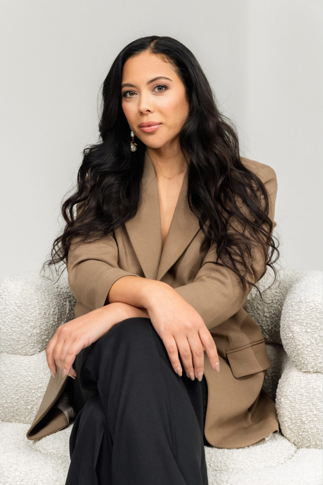 A woman weating a beige blazer and black pants sits in a chair with her arms folded and her legs crossed.